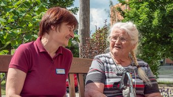 Pflegerin und Bewohnerin unterhalten sich Pflegerin unterhält sich mit Bewohnerin draußen auf der Bank.