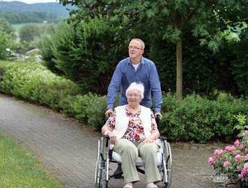 Pfleger schiebt Frau mit Rollstuhl durch Garten des Pflegeheimes.